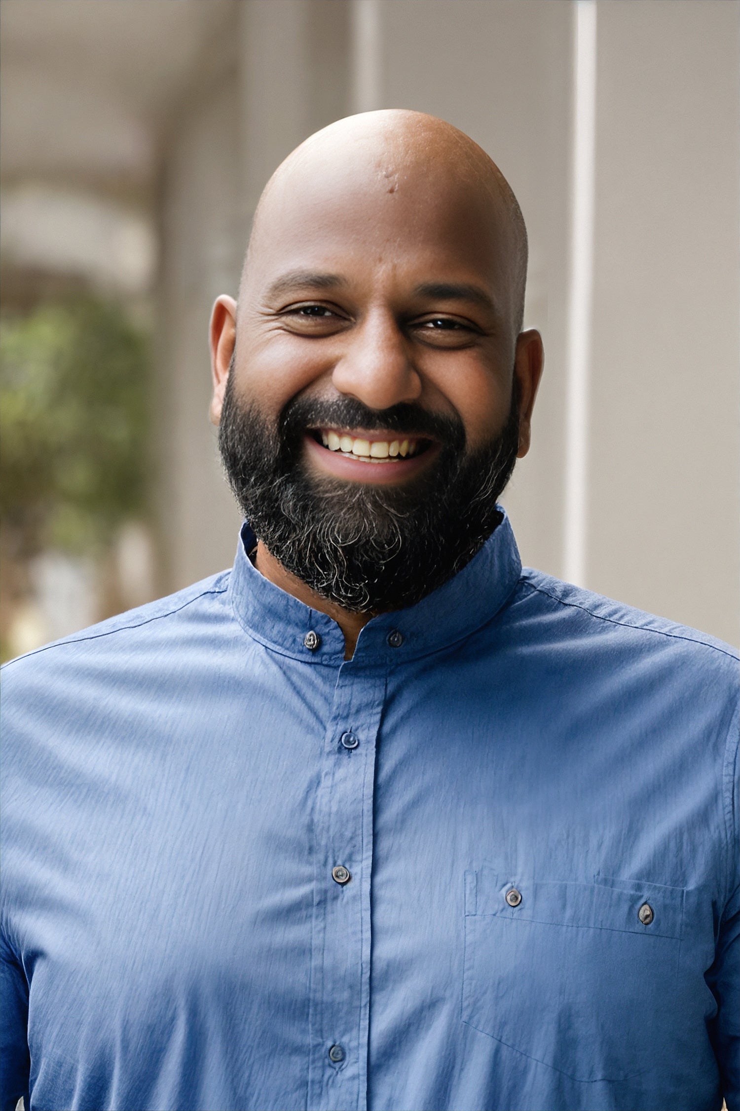 A man with a beard and mustache stands in a blue button up shirt smiling at the camera