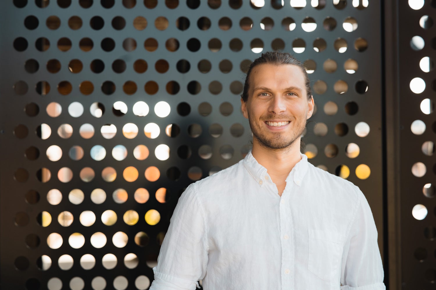 A man in a white button up shirt stands smiling in front of a metal wall