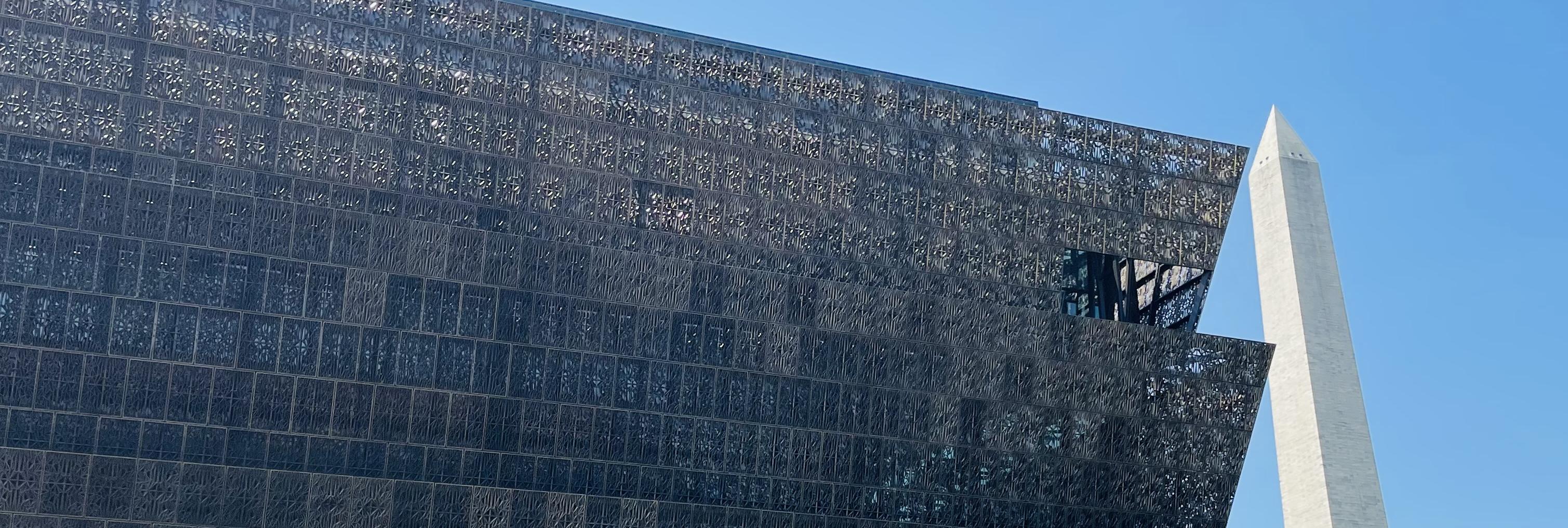  Looking up at the facade of the National Museum of African American American History and Culture with the Washington Monument in the background to the right against a clear blue sky. The NMAAHC can be described as stacked trapezoids where the edge of each level points diagonally upward and out from the center of the building. Then the next level is smaller the the top of the one below it and grows up and out in the same way. The panels of the facade are blocks and built with metal that allows you to see li