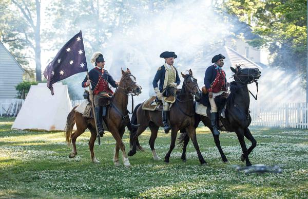 a reenactment of a Revolutionary War battle scene featuring 3 uniformed soldiers riding horses and carring a blue flag with white stars