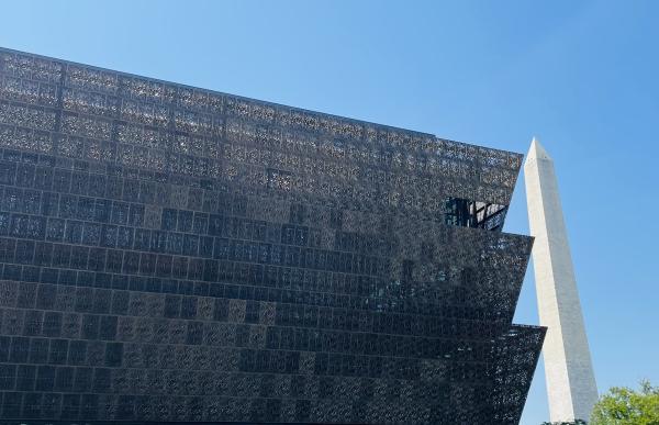  Looking up at the facade of the National Museum of African American American History and Culture with the Washington Monument in the background to the right against a clear blue sky. The NMAAHC can be described as stacked trapezoids where the edge of each level points diagonally upward and out from the center of the building. Then the next level is smaller the the top of the one below it and grows up and out in the same way. The panels of the facade are blocks and built with metal that allows you to see li